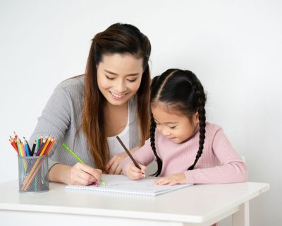 mom and daughter looking down at paper and coloring; spiral notebook; cup of different colored pencils