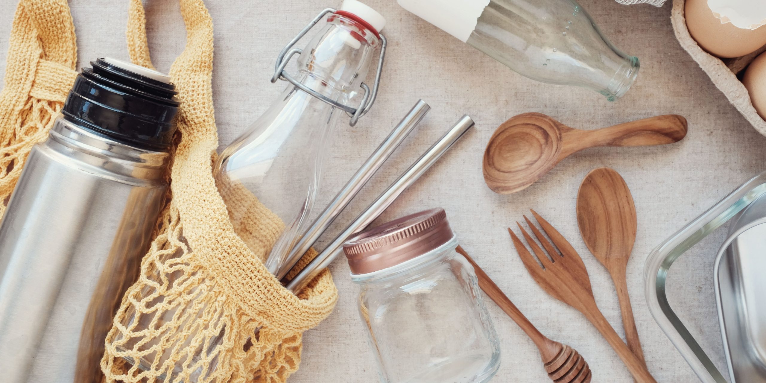 Photo shows reusable utensils, straws, a class jar, a metal thermos, and a glass bottle inside a woven bag.