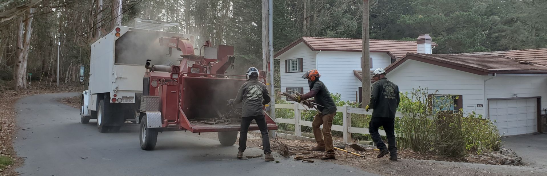 Photo shows a street and 3 people feeding wood into a wood chipper. There's a white house and trees in the background.